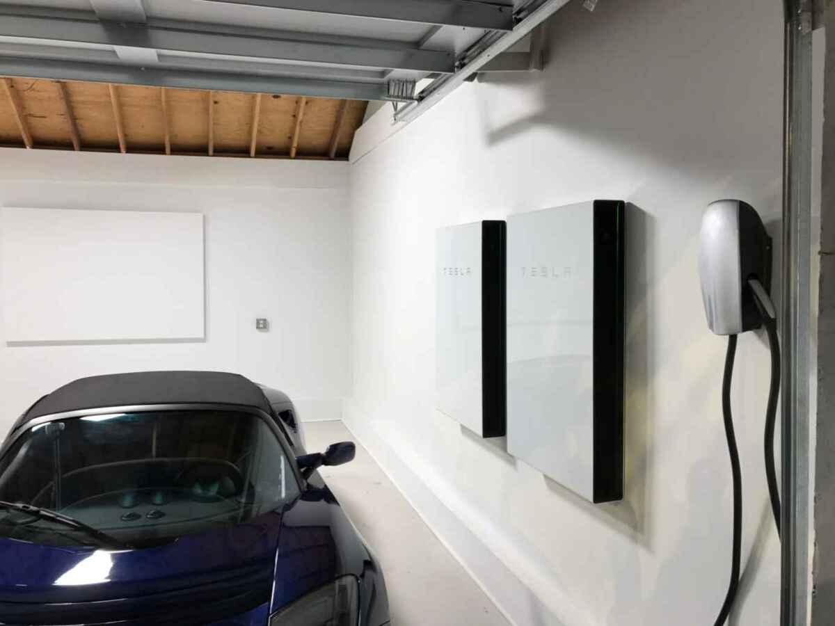 Two Tesla Powerwall units mounted on a garage wall next to an EV charger, with a Tesla Roadster parked below, showcasing home energy backup and EV integration.