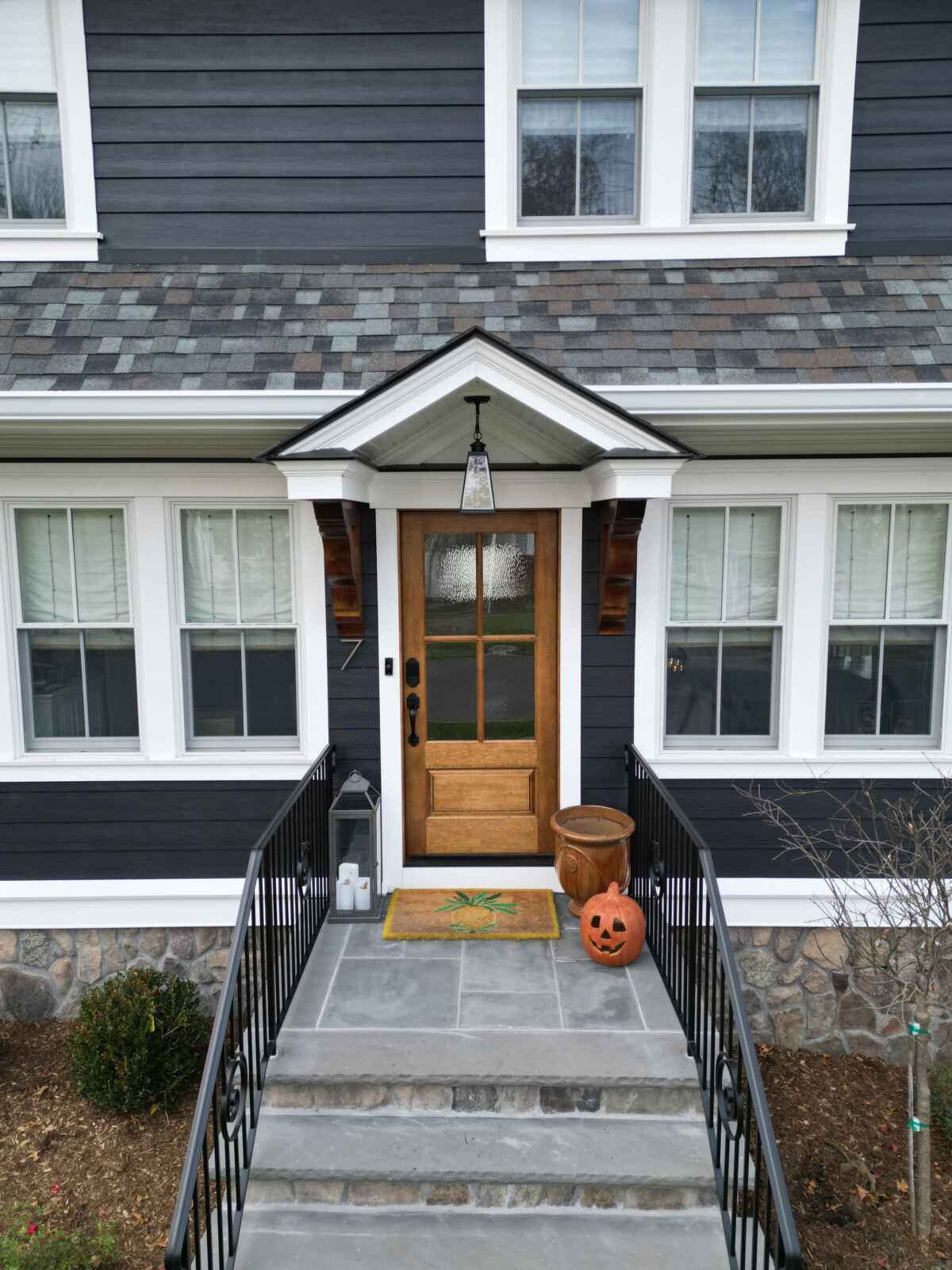 Close-up of a front entryway with dark navy horizontal siding, white trim, a natural wood front door, and seasonal fall decorations.