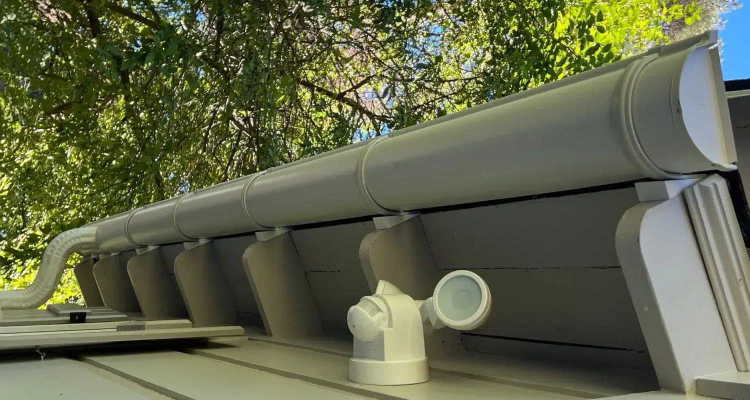 Close-up of a modern gutter system with downspout and brackets mounted on a roof edge, surrounded by leafy trees in the background.