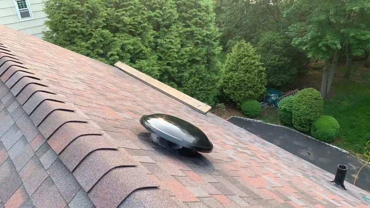 GAF Master Flow power fan installed on a red asphalt shingle roof near the ridge, surrounded by lush green trees and adjacent to a residential driveway.