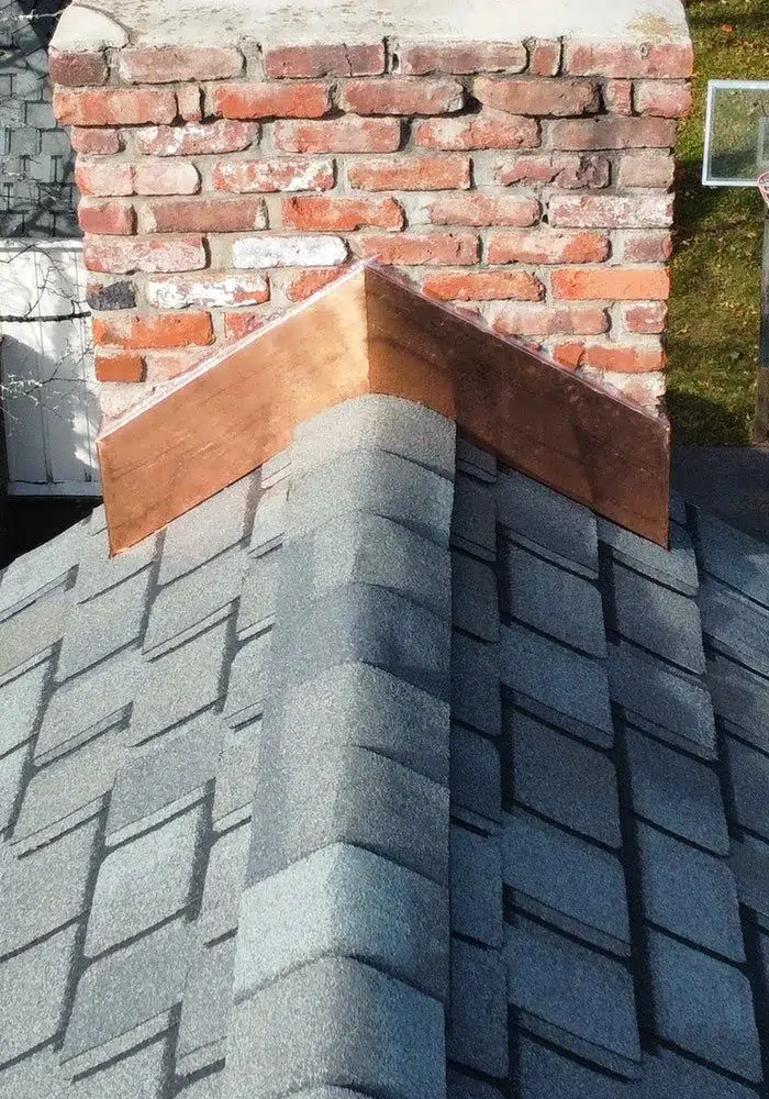 Close-up of an asphalt shingle roof with new copper flashing installed at the chimney intersection, illustrating signs of a recent roof replacement.
