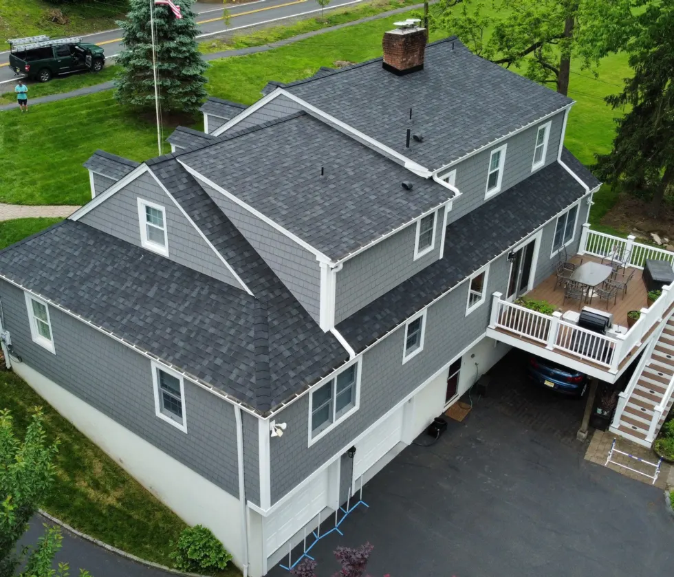 Aerial view of a large residential home with a newly repaired asphalt shingle roof, gray siding, white trim, and a second-story deck.