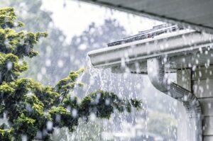Storm water is overflowing off the tile shingle roof - streaming, rushing and splashing out over the overhanging eaves trough aluminum roof gutter system on a suburban residential colonial style house near Rochester, New York State, USA during a torrential mid-summer July downpour.
